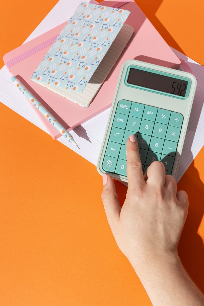 A hand pressing keys on a mint-green calculator lying on a pink notebook and graph paper against an orange background.