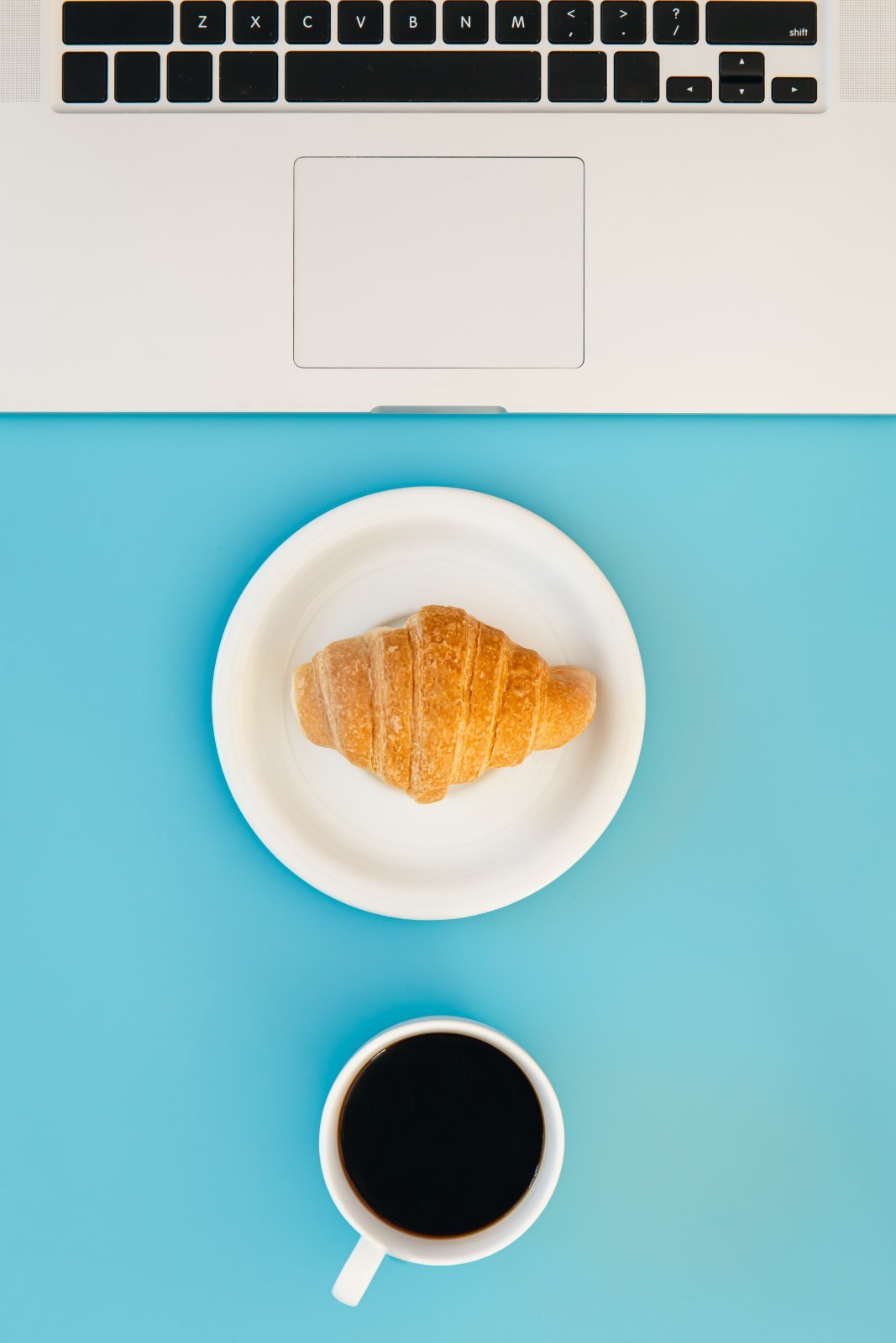 A cup of black coffee and a croissant on a white plate beside an open laptop, on a light blue background.